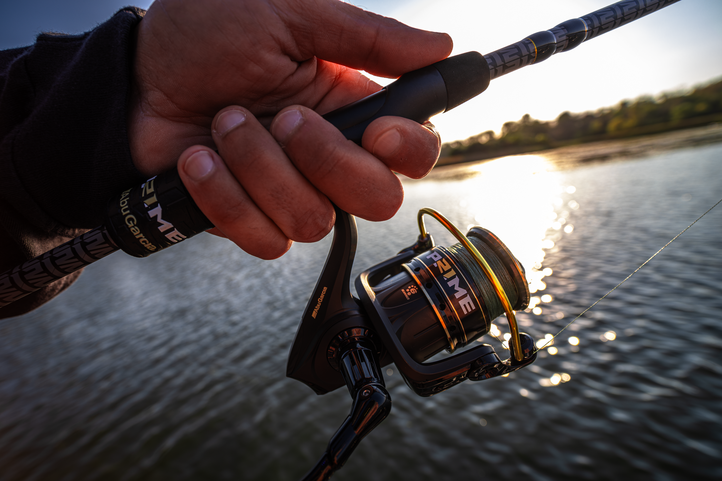 Abu Garcia + PRIME21 Collaboration Spinning Combo held by a hand over a body of water during sunset.