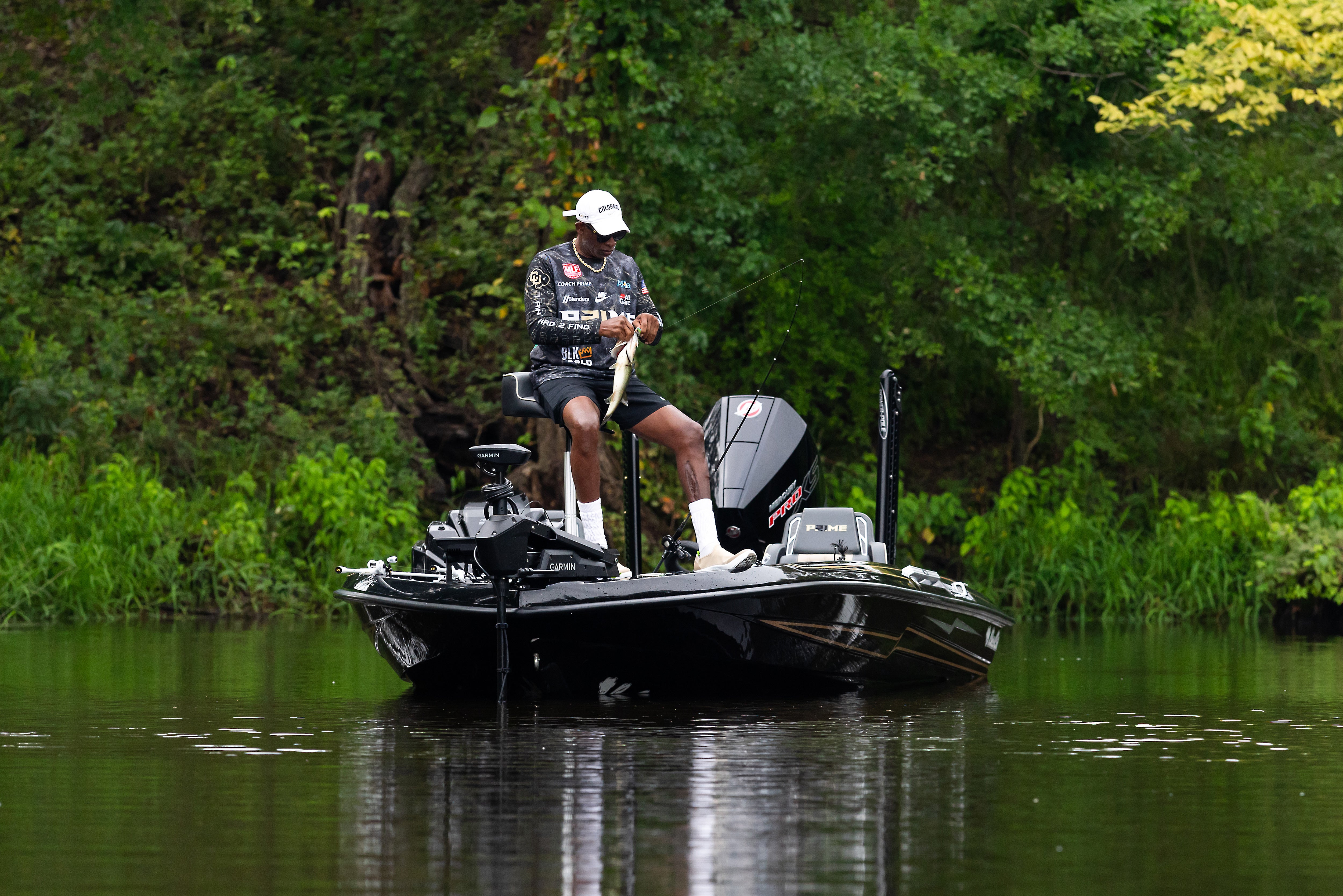 Coach Prime on a boat in a lake surrounded by trees holding a fish and a combo. 
