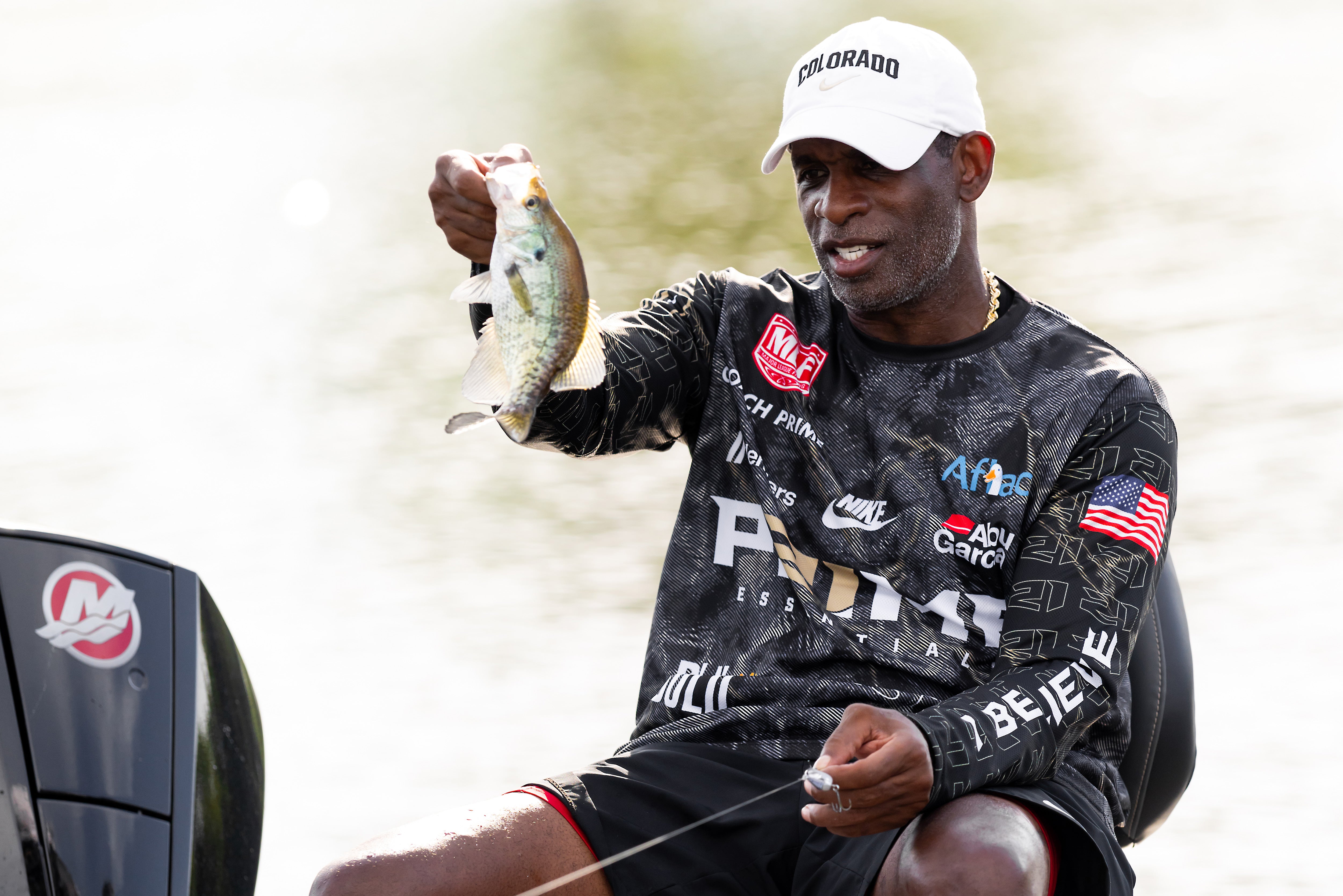 Coach Prime holding a fish while sitting on a boat, wearing a Colorado cap and a patterned shirt with various logos.