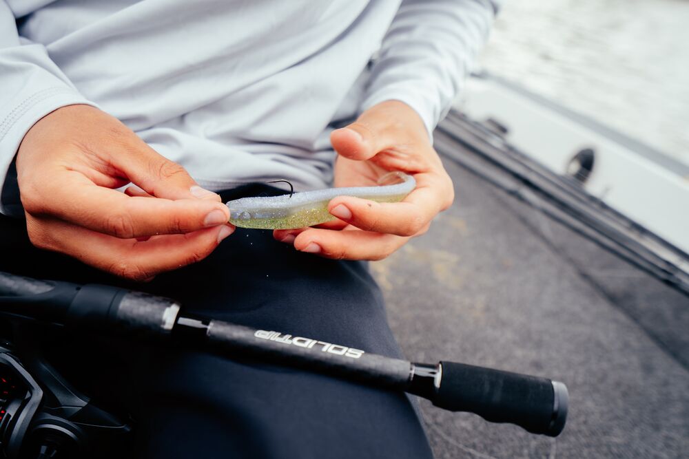 A close-up lifestyle shot of a person holding a soft fishing lure in their hands, seated on a boat with a Abu Garcia Veritas Tournament Solid Tip rod visible in the foreground.